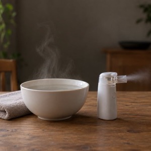 A steaming bowl with a towel beside a small handheld nebulizer giving off a fine mist